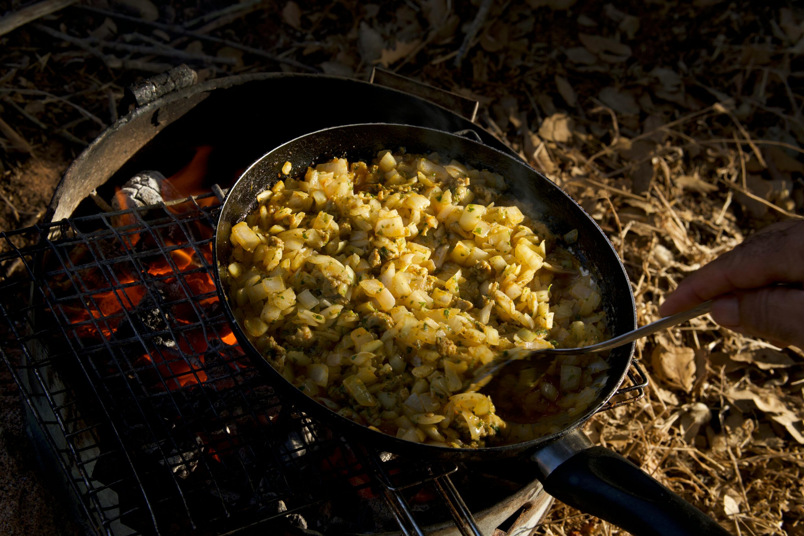 Stirring a pan of diced vegetables over a campfire grill in a rustic outdoor setting.